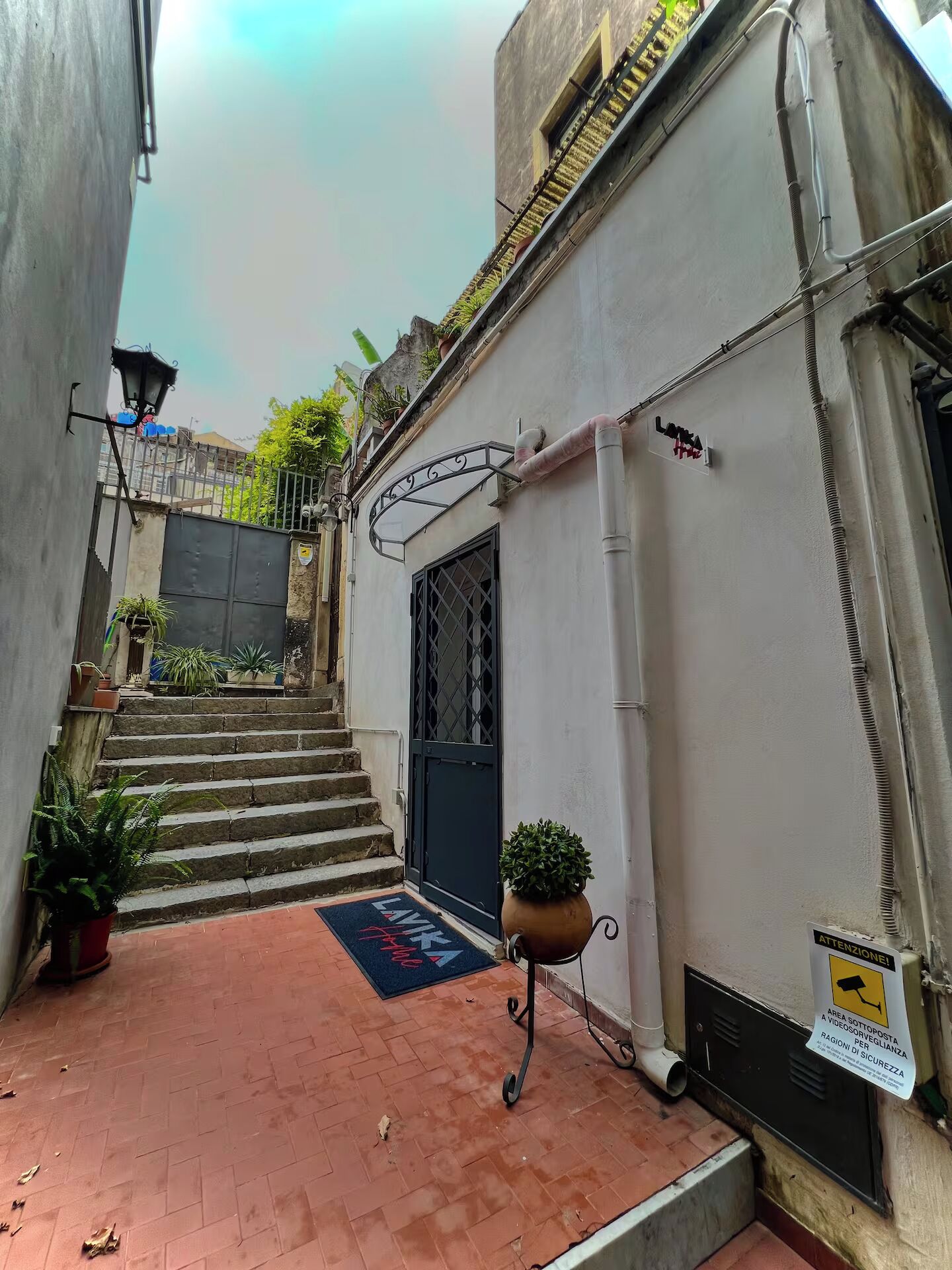 Une ruelle étroite avec un portail noir, un paillasson rouge sur lequel est écrit « LAVAKIA », et une plante en pot.