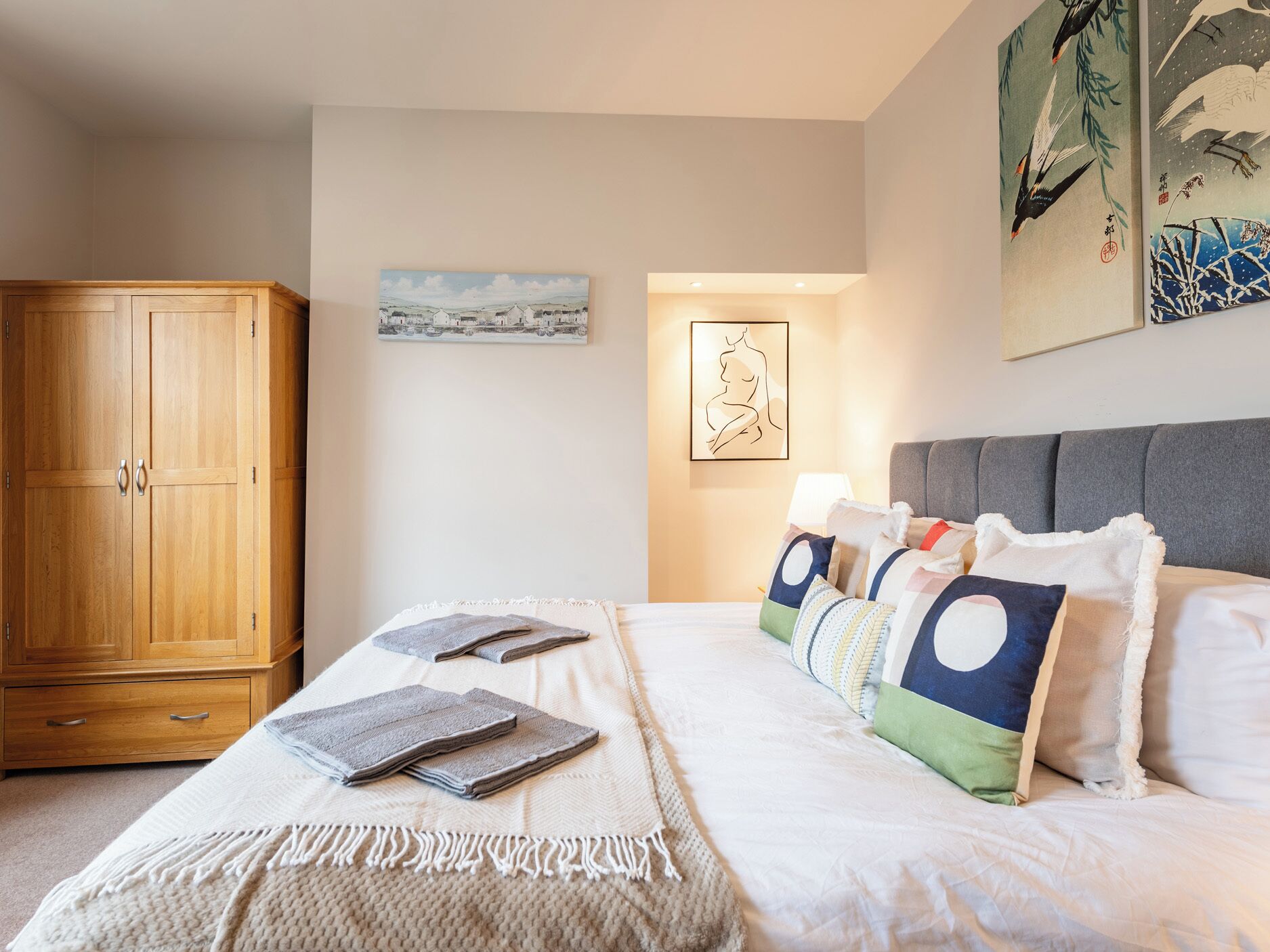 A bedroom with a bed, pillows, a blanket, a wooden wardrobe, and wall art.