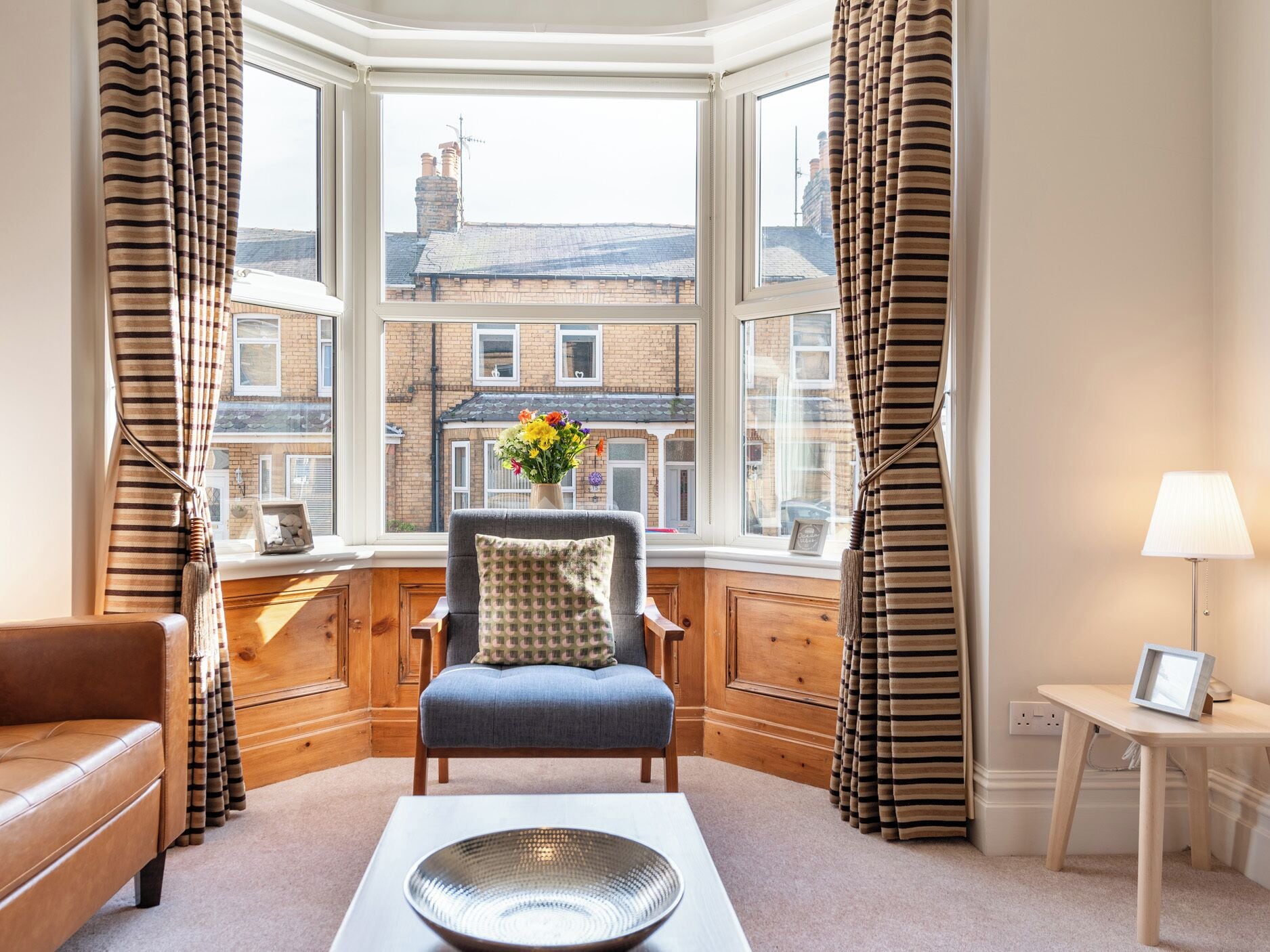 A corner room with a bay window, a sofa, a chair, a table with a bowl, and curtains.