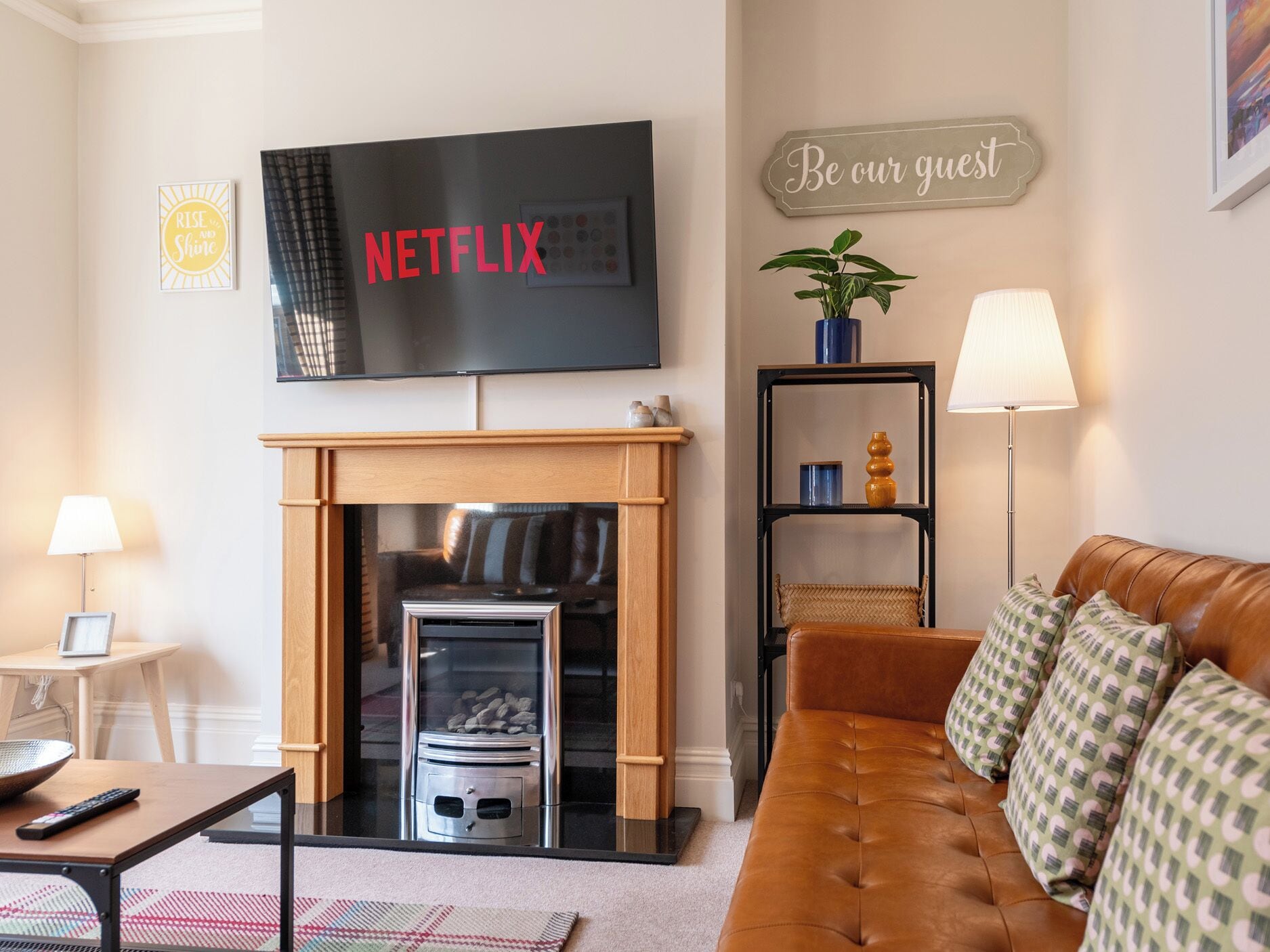 Una sala de estar con chimenea, televisor montado encima, sofá y un cartel en la pared que dice 'Be our guest'.