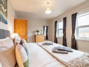 Une chambre à coucher comprenant un lit, des oreillers, une commode, un miroir, une fenêtre avec des rideaux et un luminaire suspendu.