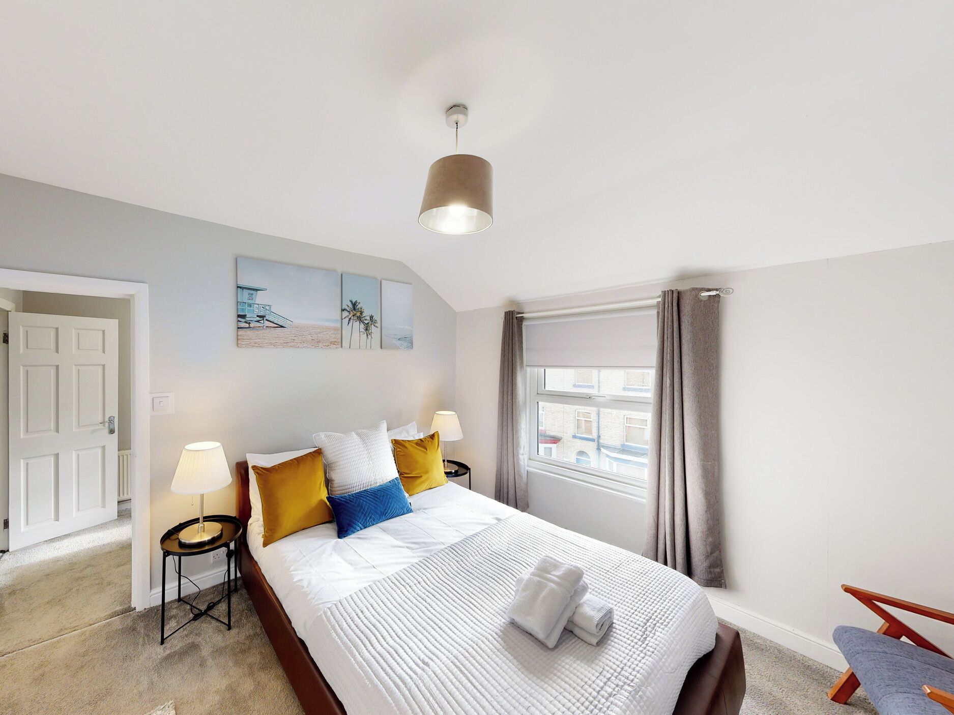 A hotel room with a bed, bedside lamps, a window with curtains, a door, and artwork on the wall.