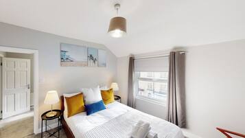A hotel room with a bed, bedside lamps, a window with curtains, a door, and artwork on the wall.