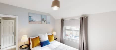 A hotel room with a bed, bedside lamps, a window with curtains, a door, and artwork on the wall.