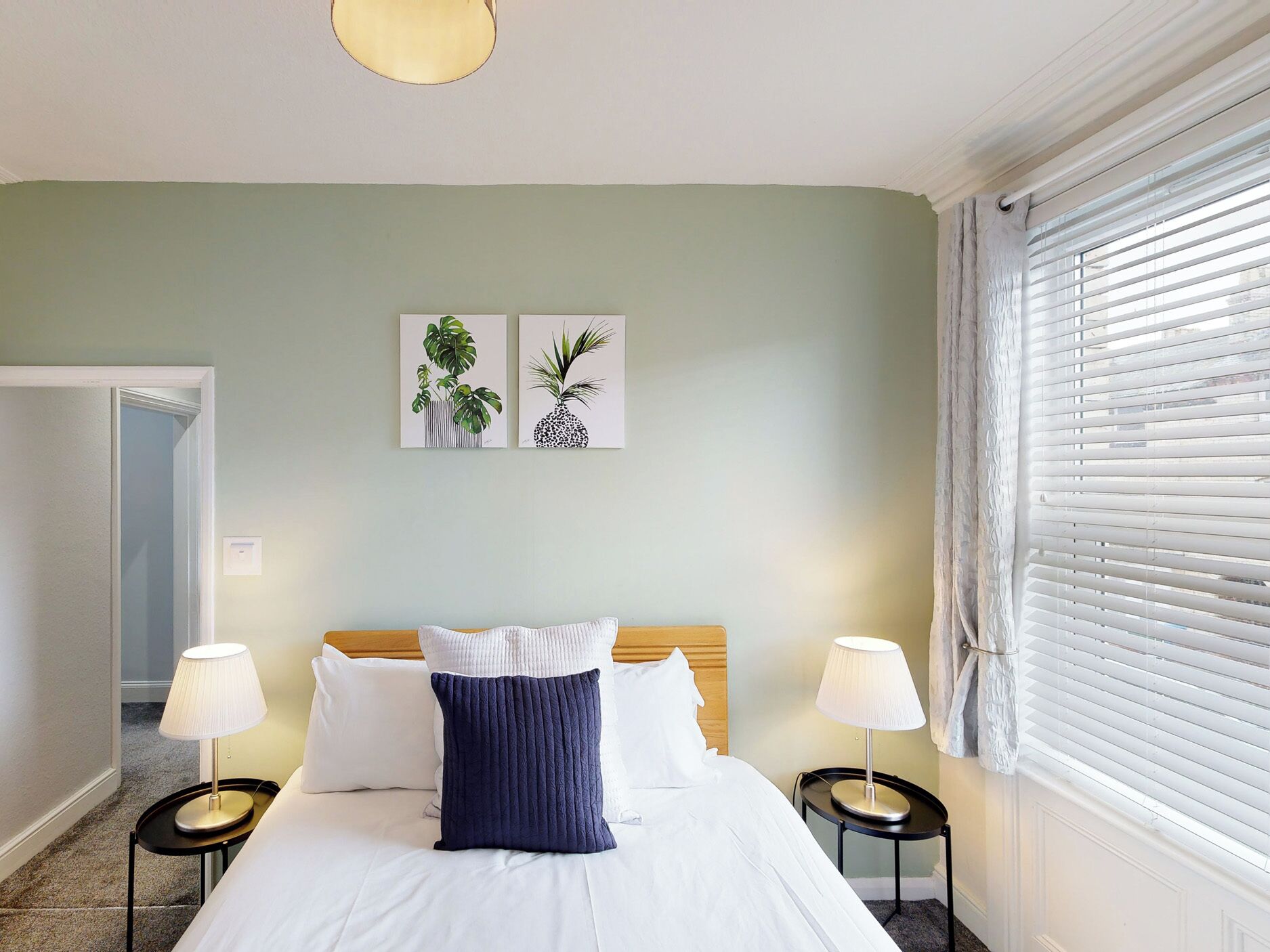 Habitación de hotel con cama, dos mesitas de noche, una ventana con persianas y dos cuadros colgados en la pared.