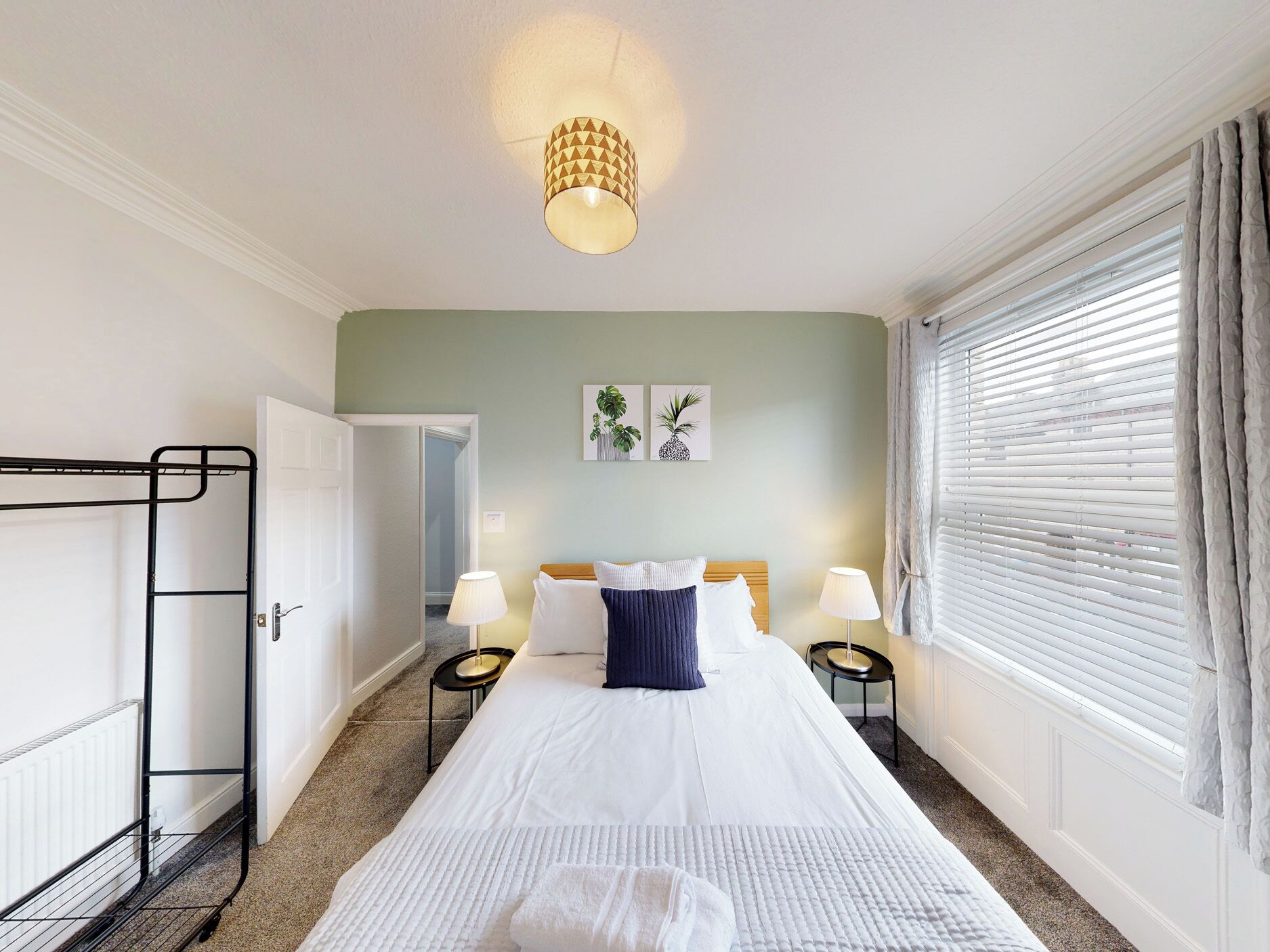 Habitación de hotel con cama, dos lámparas de noche, aplique de pared, ventana con persianas y puerta que da a otra habitación.