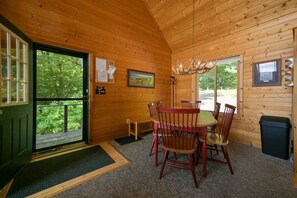 Ein Interieur einer Holzhütte mit Esstisch und Stühlen, einer Bank, einer Chandelüster und einer zur Außenwelt führenden Tür.
