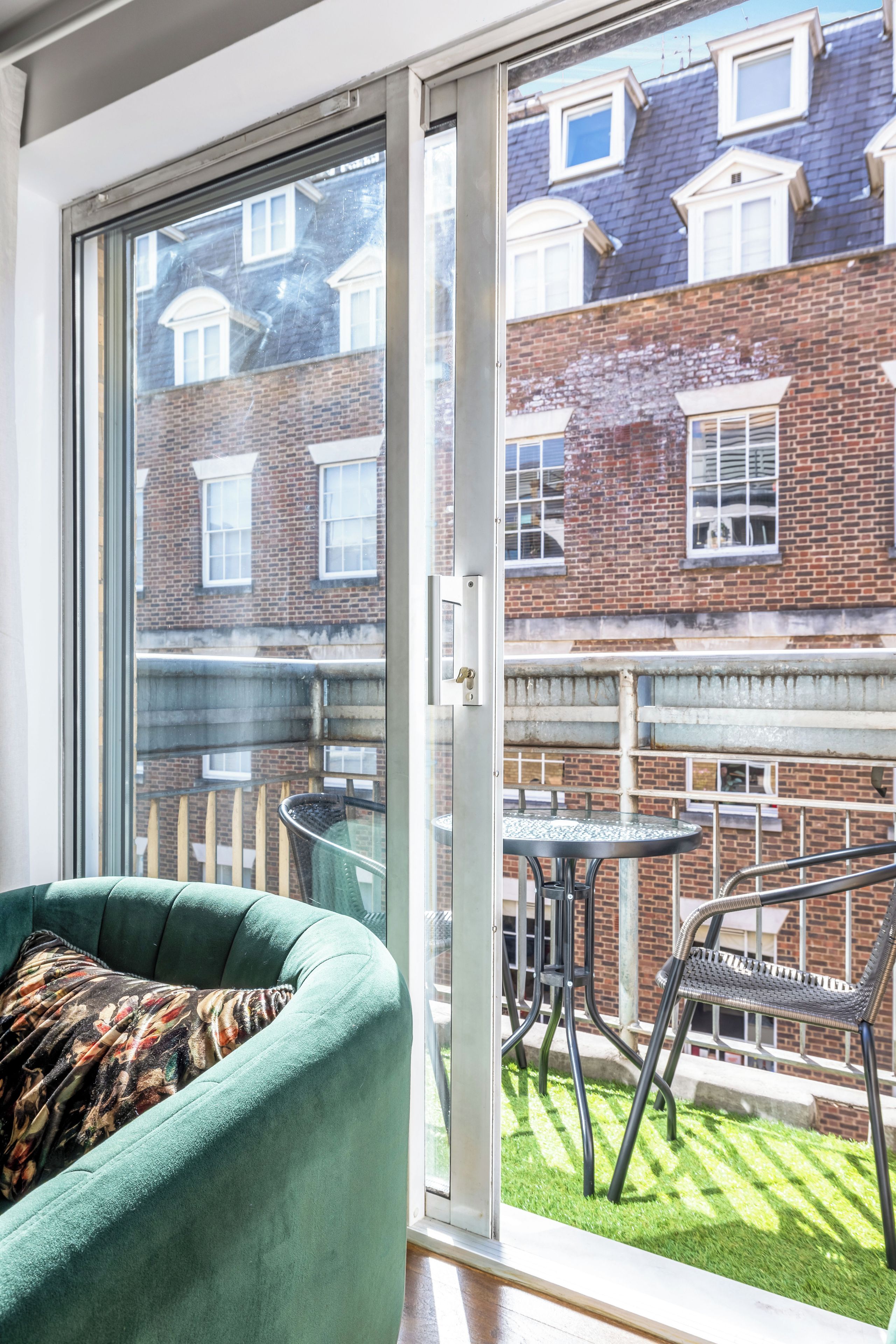 A room with a sliding glass door leading to a balcony with a table and chairs, a green armchair, and a view of a brick building with windows.