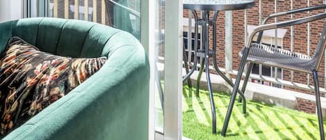A room with a sliding glass door leading to a balcony with a table and chairs, a green armchair, and a view of a brick building with windows.