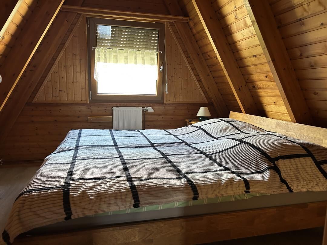 Ein Zimmer mit schrägverdeckter Decke, einem Fenster mit Jalousien, einem Bett mit gemustertem Überwurf und einem Heizkörper.