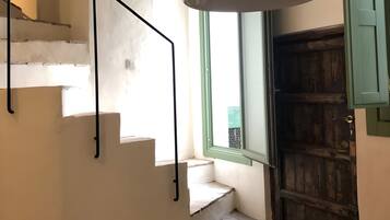 Una escalera con barandilla negra, una lámpara colgante y una puerta de madera con contraventanas verdes.