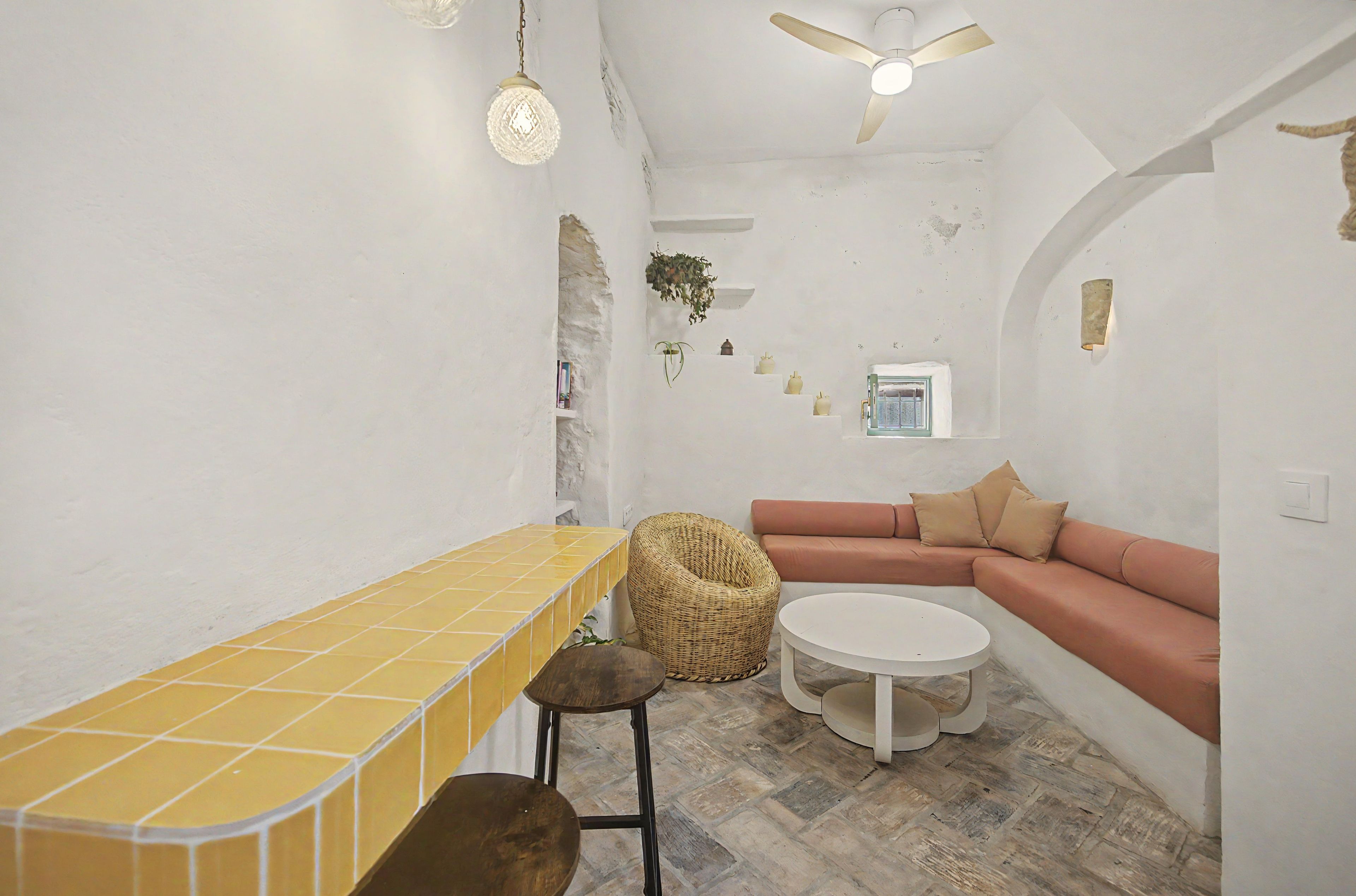 Una habitación con un banco de azulejos amarillos, una mesa redonda blanca, una silla de mimbre marrón, un sofá rojo con cojines beige y un ventilador de techo.