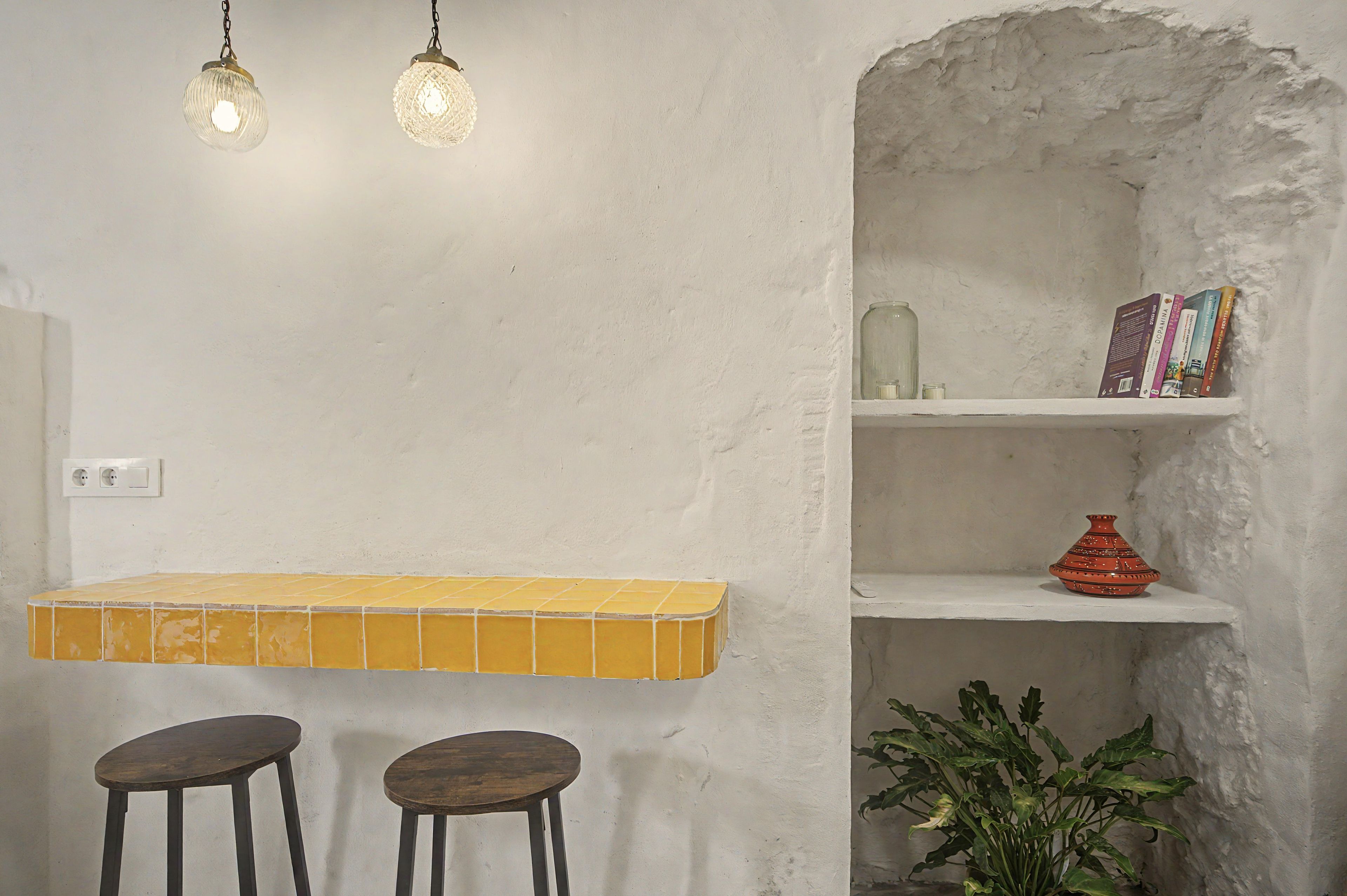 A room with a yellow tiled bar counter, two stools, a shelf with books and a vase, and a hanging light fixture.