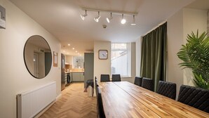 Un comedor con mesa y sillas de madera, un espejo redondo, un reloj y una ventana con persianas.
