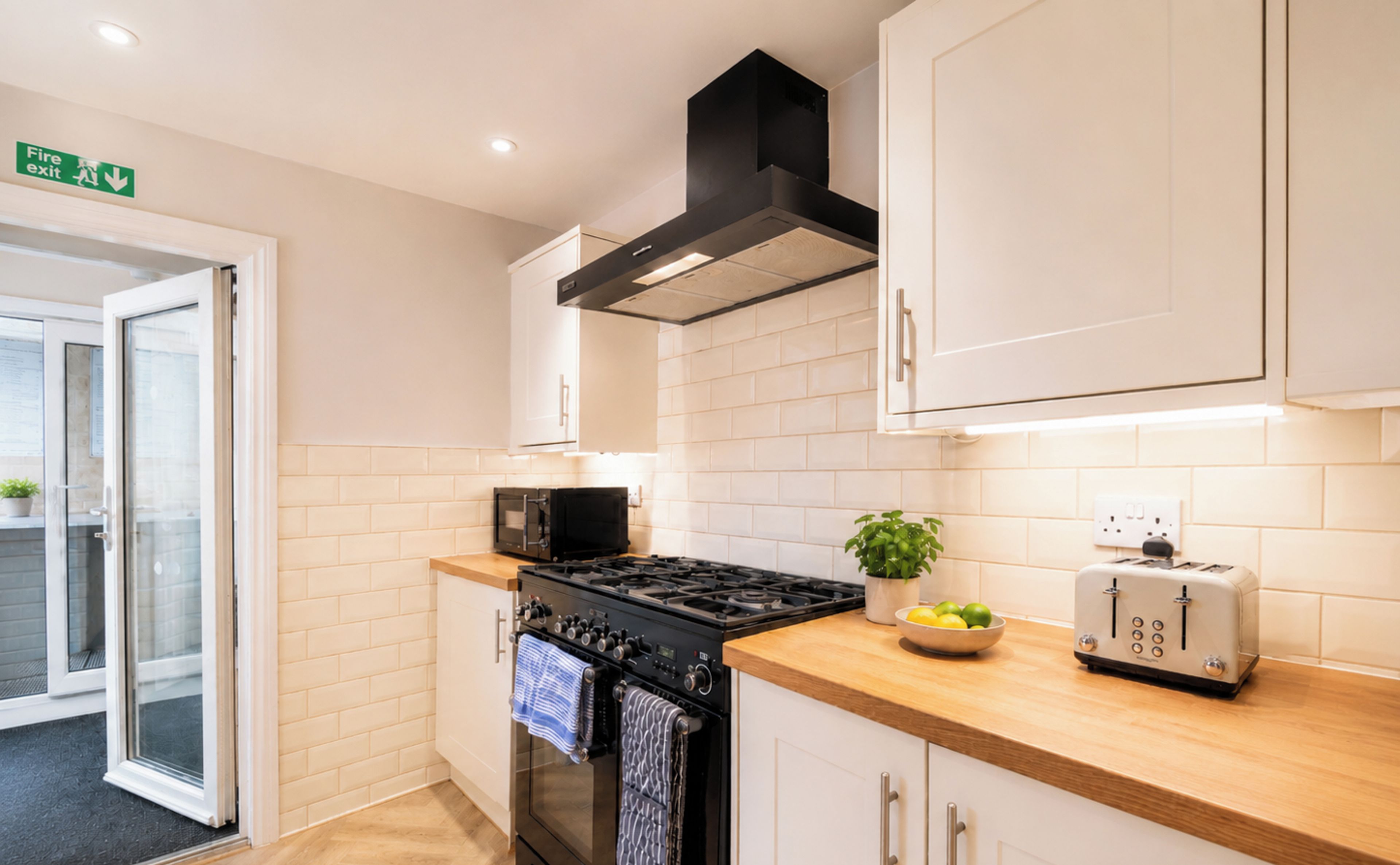 A modern kitchen with a stove, microwave, and toaster. There is a fire exit sign above the door.
