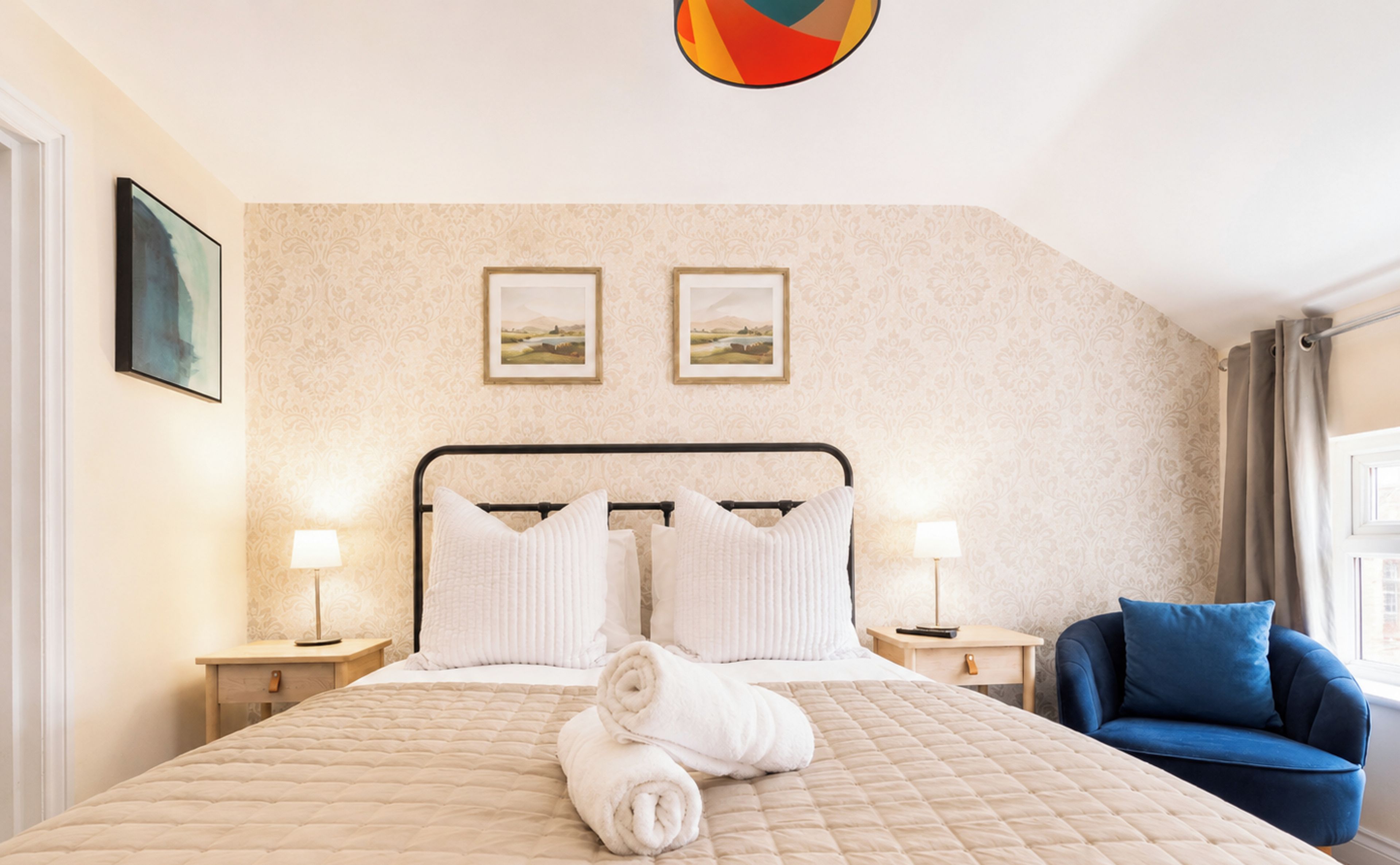 A hotel room with a bed, two bedside lamps, a blue armchair, and two framed pictures on the wall.