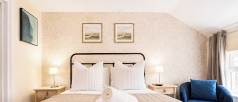A hotel room with a bed, two bedside lamps, a blue armchair, and two framed pictures on the wall.