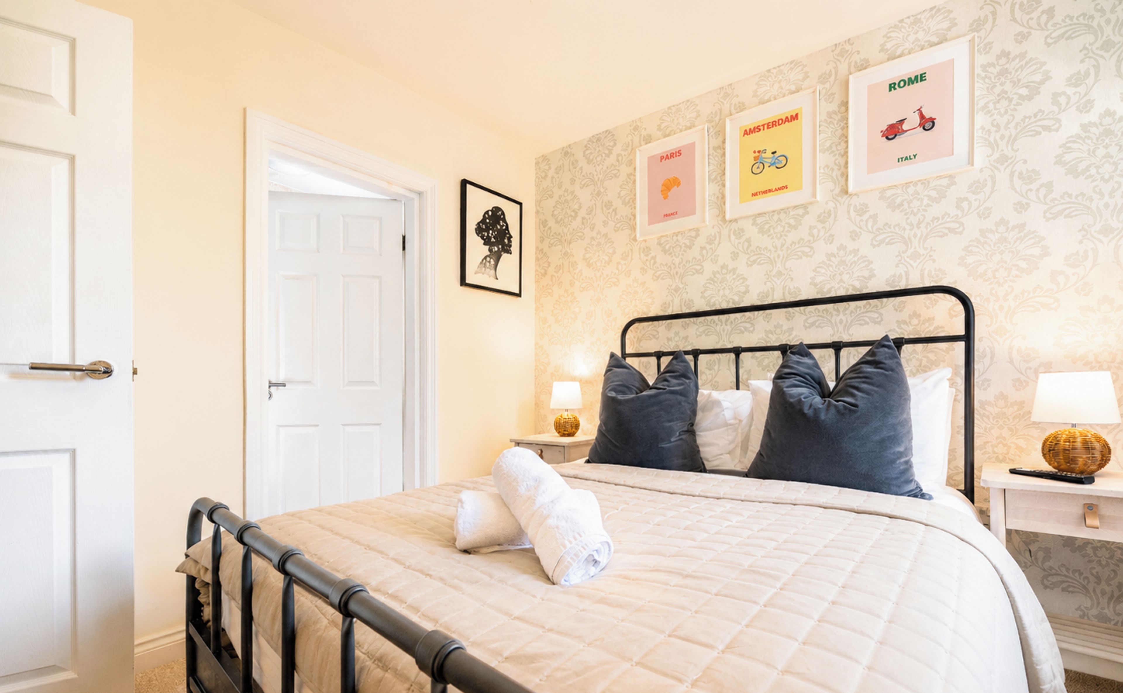 A bedroom with a bed, a door, a wall with framed pictures, and a nightstand with a lamp.