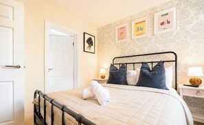 Ein Schlafzimmer mit einem Bett, einer Tür, einer Wand mit gerahmten Bildern und einem Nachttisch mit einer Lampe.