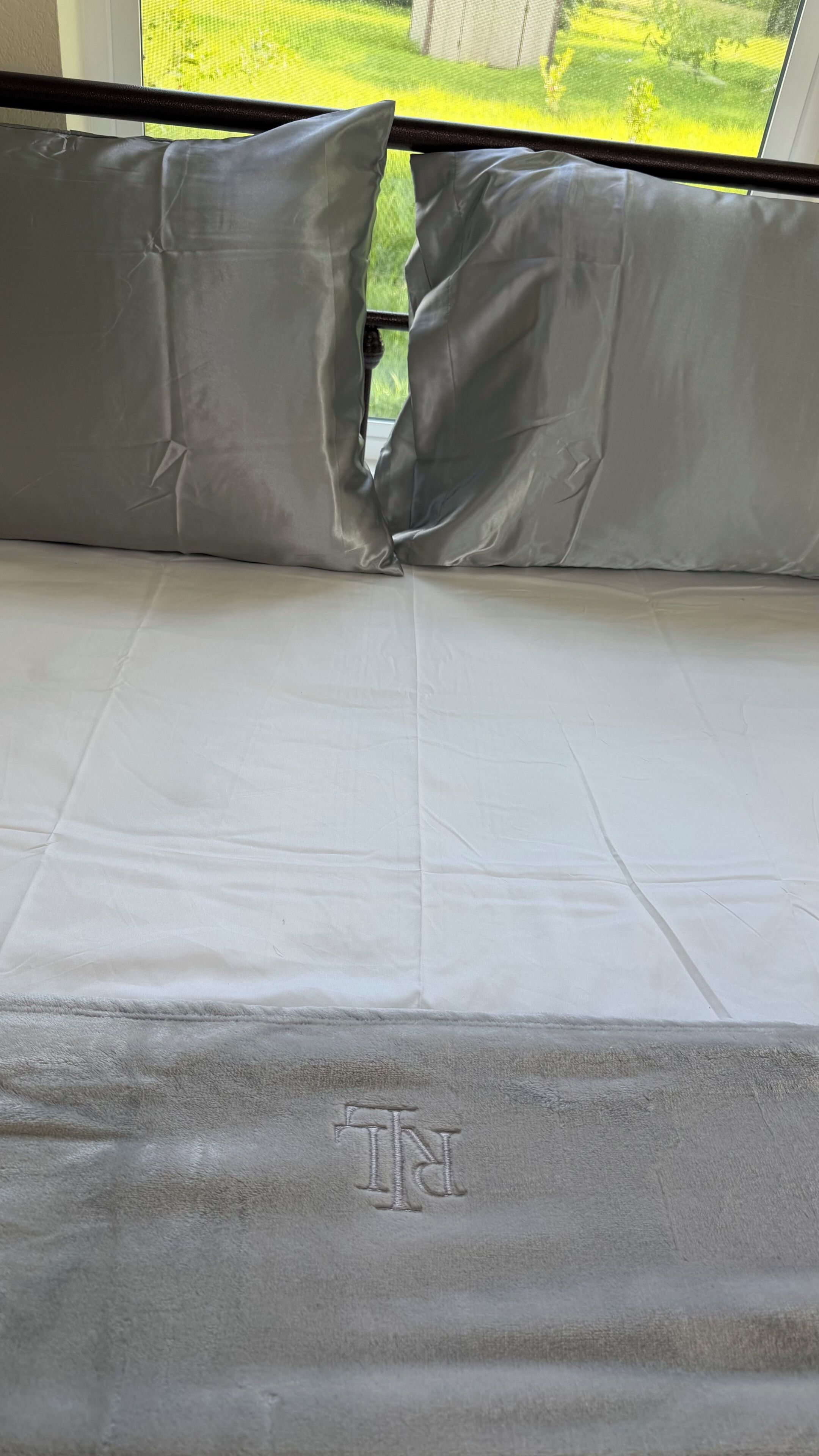 A bed with a white mattress and grey pillows, a grey blanket with a monogram, and a window view of a green landscape.