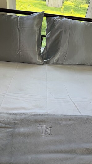 Uma cama com colchão branco e almofadas cinzas, um cobertor cinza com monograma, e vista para uma paisagem verde pela janela.
