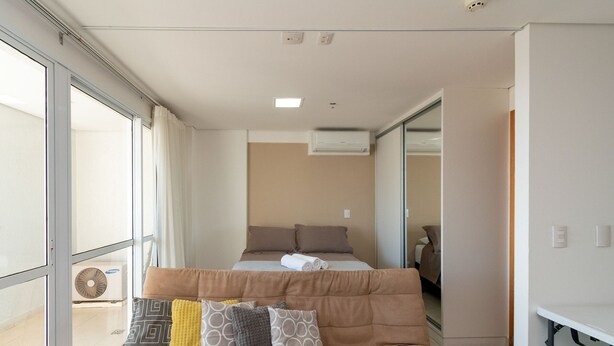 A hotel room with a bed, a window, a mirror, and an air conditioning unit. - Flat Near Flamboyant Mall - Bft3207 (Goiânia)
