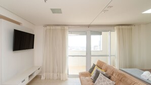A modern living room with a flat-screen TV, a sofa with patterned cushions, and a sliding glass door leading to a balcony. - Flat Near Flamboyant Mall - Bft3207 (Goiânia)