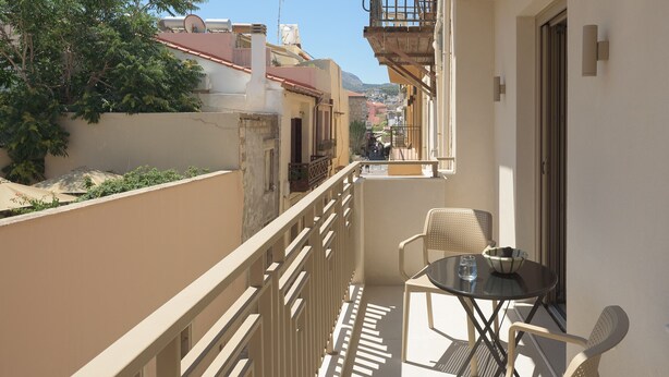 En balkon med et bord og stole, der giver udsigt over en gade med bygninger og et fjernt beliggende bjerg.