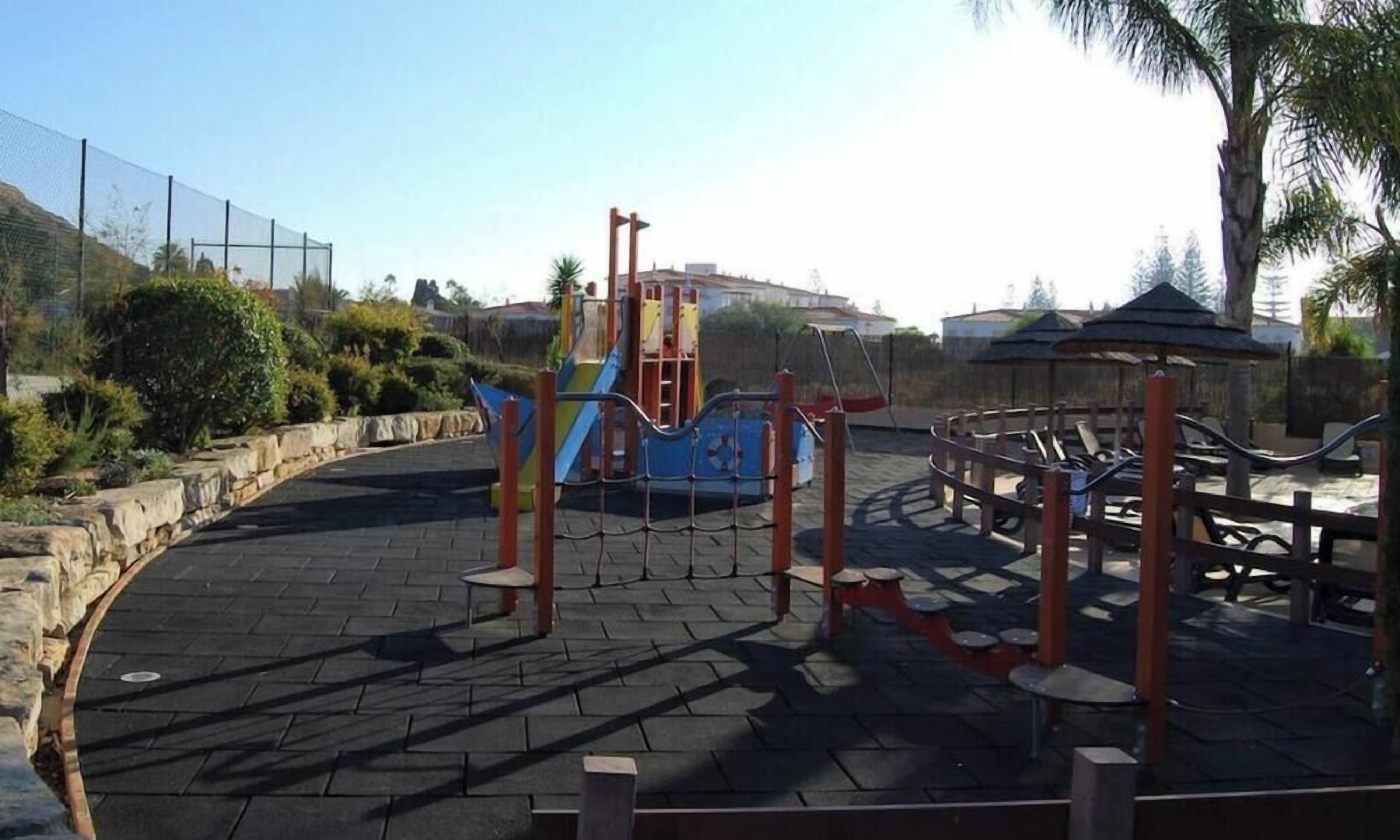 Un parque infantil con una estructura para escalar, un tobogán y un área cercada con bancos.