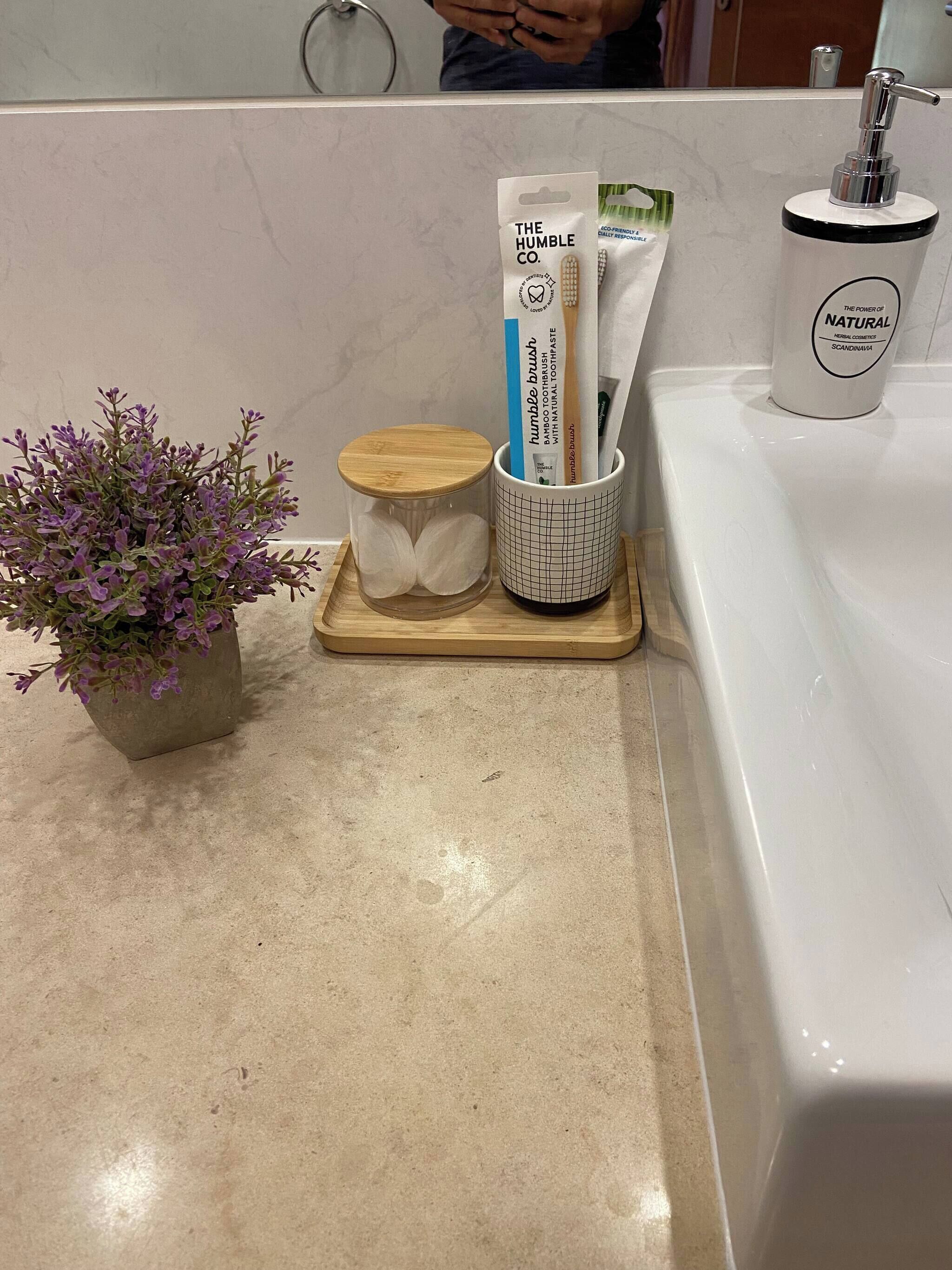 A bathroom counter with a potted plant, a wooden container, a mug with a grid pattern, and a bottle of natural herbal cosmetics.