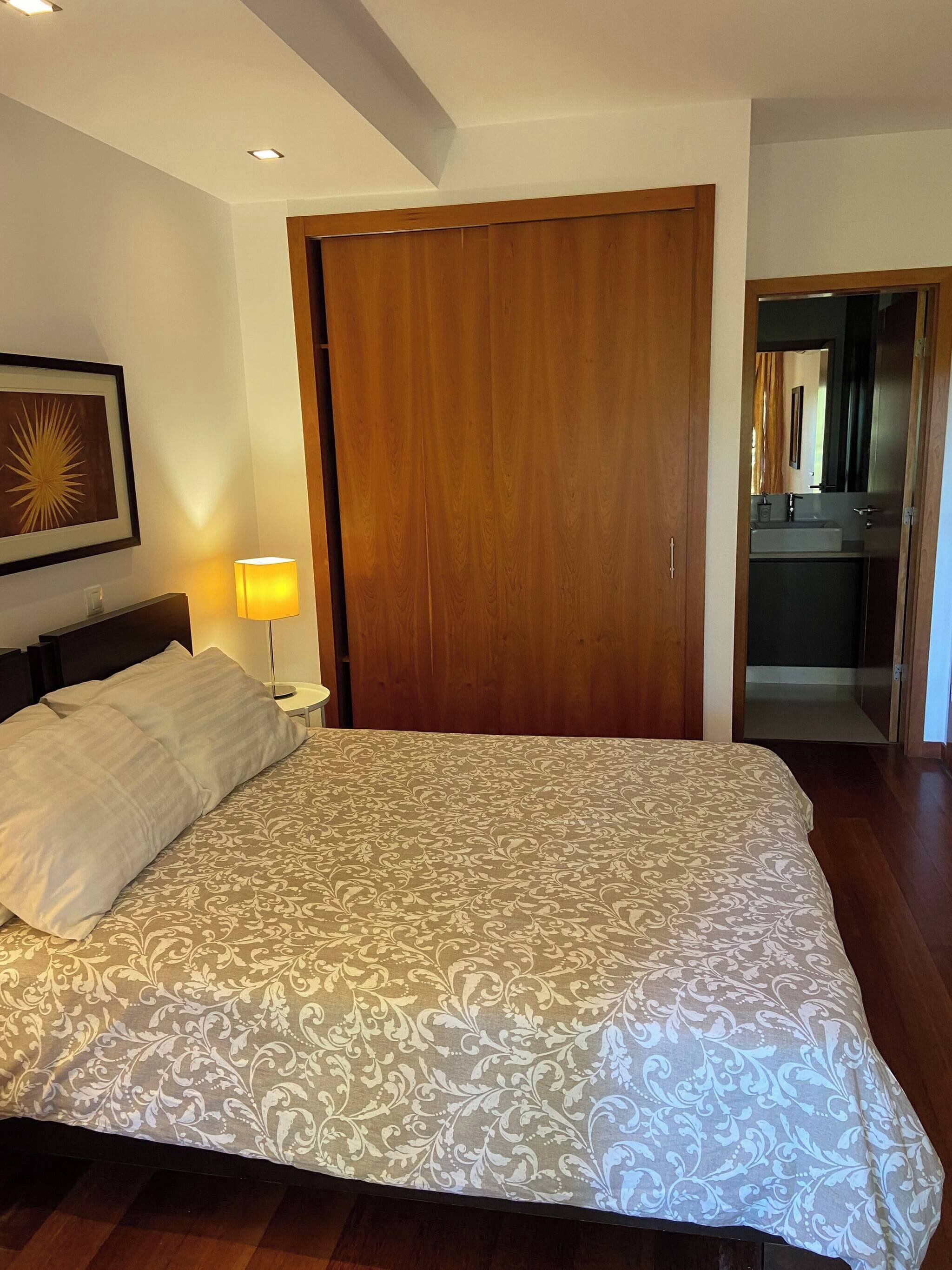 Ein Schlafzimmer mit einem großen Bett, einem Holzschrank, einer Nachttischlampe und einem gerahmten Bild an der Wand.