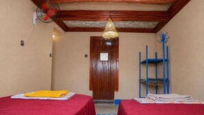 Una habitación con dos camas, una puerta de madera, un estante azul y un ventilador.