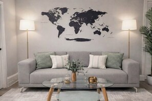 Ein Wohnzimmer mit grauem Sofa, zwei Lampen, einer Weltkarte an der Wand und einem Glastisch mit einer Vase.