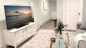 A living room with a television on a white cabinet, a glass coffee table, a grey sofa, and a door in the background. - Comfortable English Appartment Walk to A Union Market (Washington)