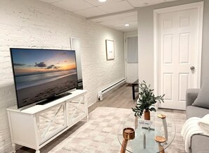 Ein Wohnzimmer mit Fernseher auf einem weißen Schrank, einem Glastisch, einem grauen Sofa und einer Tür im Hintergrund.