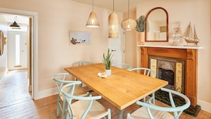 Een eetkamer met een houten tafel, lichtblauwe stoelen, hangende rieten lampen, een open haard en een spiegel erboven.