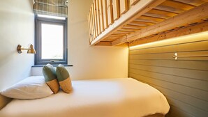 Een enkel bed met een houten hoofdbord, een raam met zonwering, en een aan de muur bevestigde lamp.