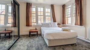 A hotel room with a large bed, a small table, a mirror, and a window with curtains. - Host Stay - Abbey House Sea View (Worthing)