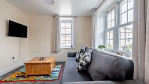 A modern living room with a sofa, a coffee table, a TV, and large windows. - Host Stay - Abbey House Sea View (Worthing)