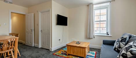 Un salon avec un téléviseur à écran plat fixé au mur, une table basse en bois, un canapé gris et un tapis coloré.