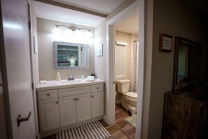 Ein Badezimmer mit einem weißen Waschbecken, Spiegel und WC. Ein Duschvorhang und ein kleiner Hocker befinden sich in der Nähe des Toilettensitzes.