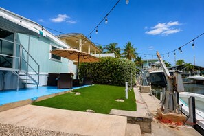 Uma marina com barcos atracados, uma área gramada com móveis de exterior e um edifício com fachada azul.