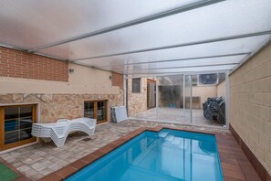 A covered outdoor swimming pool area with a stone wall, a glass door leading to a patio, and a stack of white chairs. - La Casona De Cuéllar: Rural House for Groups with Pool in Segovia (Cuéllar)