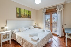 A bedroom with a large bed, a bedside table with a lamp, a painting on the wall, and a view through the window. - La Casona De Cuéllar: Rural House for Groups with Pool in Segovia (Cuéllar)