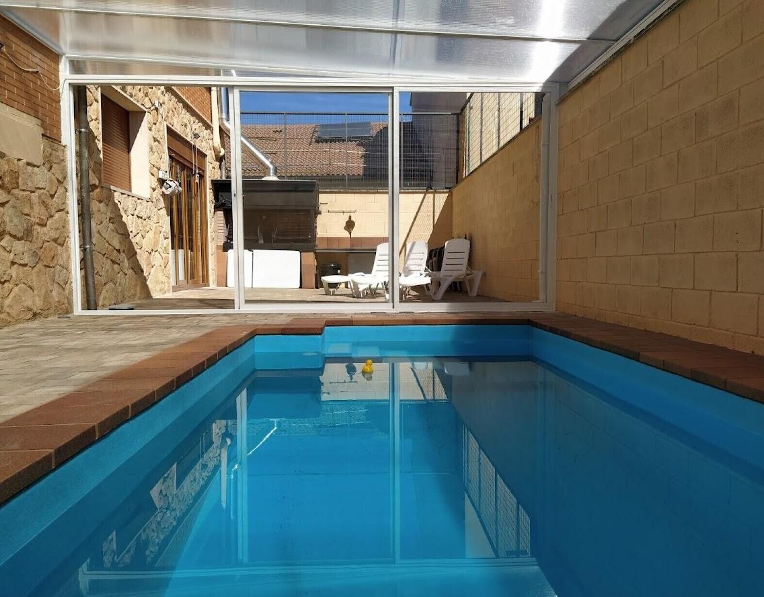 Una piscina con agua azul cristalina, rodeada por un muro de piedra y un cerramiento de vidrio.