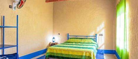 A room with a bed, a blue shelf, a patterned rug, and a window with green curtains.