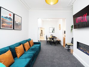 Una sala de estar moderna con un sofá color turquesa, un comedor con una mesa y sillas, y un televisor en la pared mostrando 'NETFLIX'.