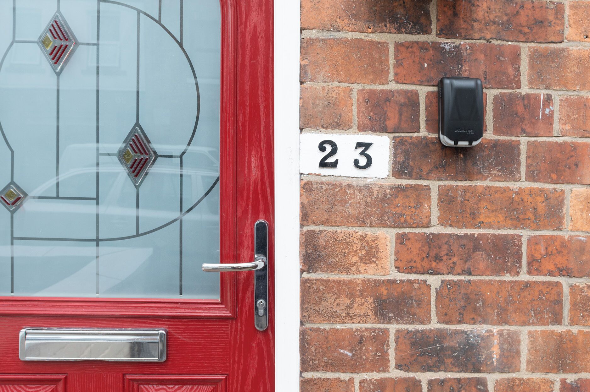 A red door with a glass panel and a mailbox, and a brick wall with a number '23' sign.