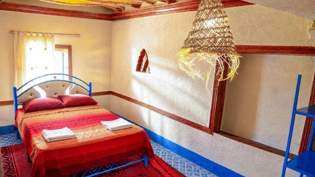 Een slaapkamer met een bed, een raam met gordijnen, een hanglamp en een kamerkleed met dessin.