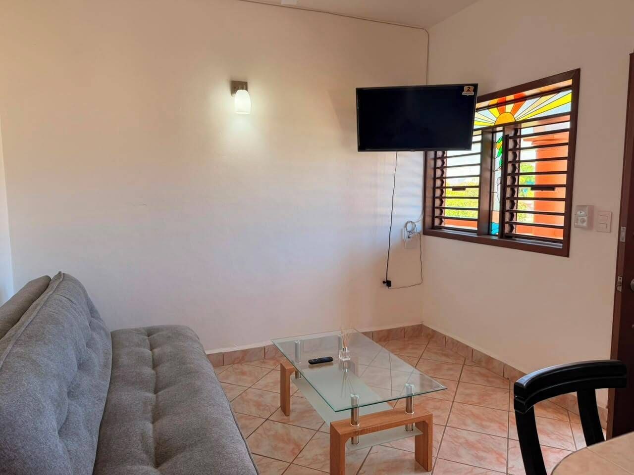 Una sala de estar con sofá, una mesa de centro de vidrio, un televisor de pantalla plana montado en la pared y una ventana con un celosía decorativa.